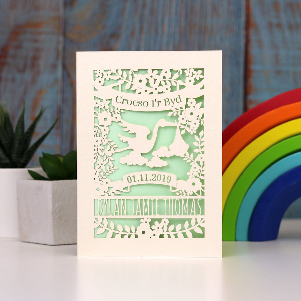 Gender neutral new baby personalised papercut card in cream with a light green insert. Shows Croeso i'r Byd , welcome to the world in Welsh, with a lovely laser cut scene showing a stork carrying a baby bundle surrounded with flowers and leaves. Space to put baby's name and date of birth under the stork.