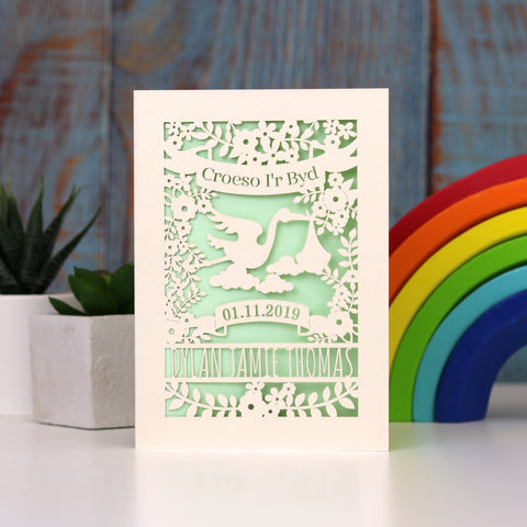 Gender neutral new baby personalised papercut card in cream with a light green insert. Shows Croeso i'r Byd , welcome to the world in Welsh, with a lovely laser cut scene showing a stork carrying a baby bundle surrounded with flowers and leaves. Space to put baby's name and date of birth under the stork. - A6 (small) / Light Green