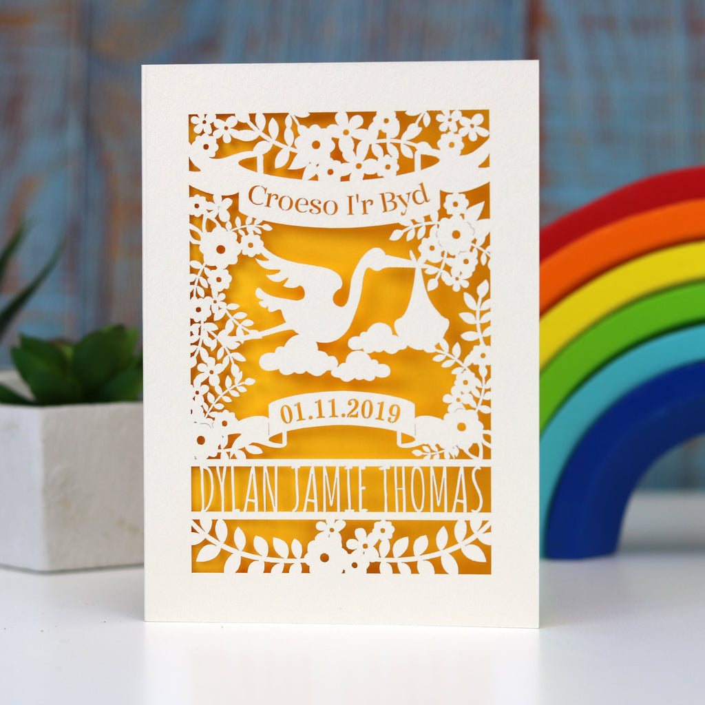 Welsh language Welcome to the World new baby papercut card. Has Croeso i'r Byd in a banner over a sweet stork bringing a baby. Flowers, leaves and space for the baby's name and date of birth. Shown here cut from cream card and finished with a  gender neutral sunshine yellow