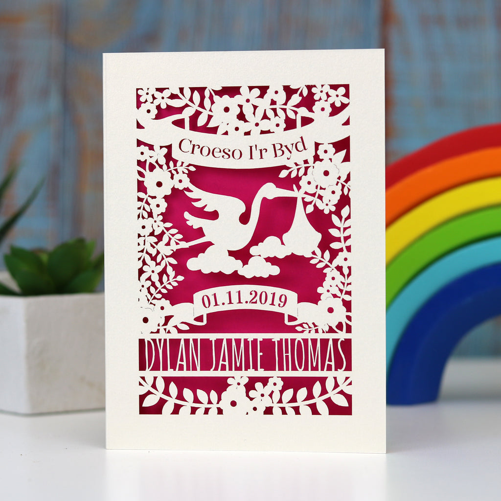 Croeso i'r Byd,  Welsh new baby personalised welcome to the world card.  Shows a lovely scene with a sweet stork carrying a baby bundle, surrounded with flowers and leaves.  Has space for baby's name and date of birth. Laser cut from cream card and finished with a bright pink insert.
