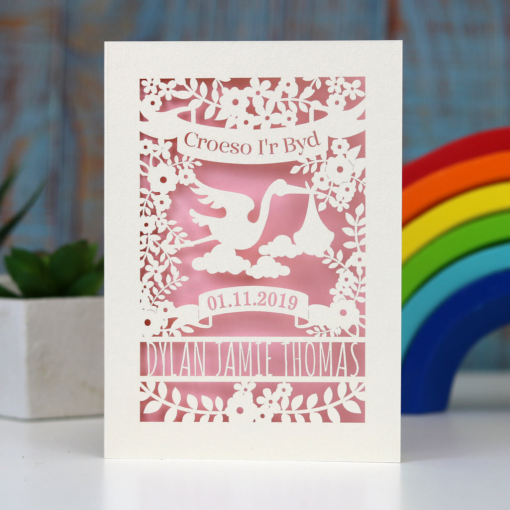 Pretty pink Croeso i'r Byd, Welsh Welcome to the world new baby papercut card.  Showing a stork, leaves and flowers with a banner for the date of birth and lines for the baby's name. Laser cut from cream card with a baby pink insert paper.