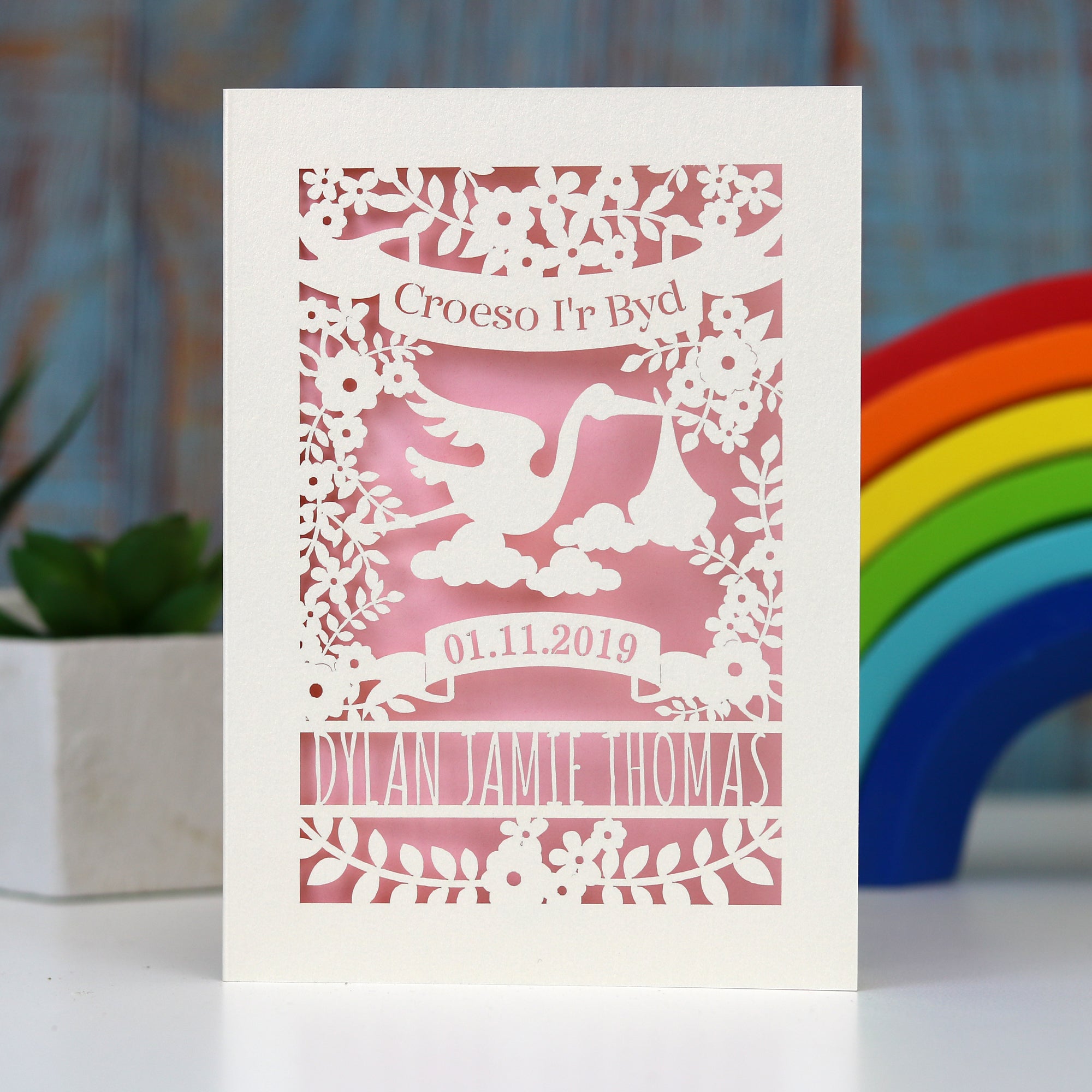 Pretty pink Croeso i'r Byd, Welsh Welcome to the world new baby papercut card.  Showing a stork, leaves and flowers with a banner for the date of birth and lines for the baby's name. Laser cut from cream card with a baby pink insert paper.