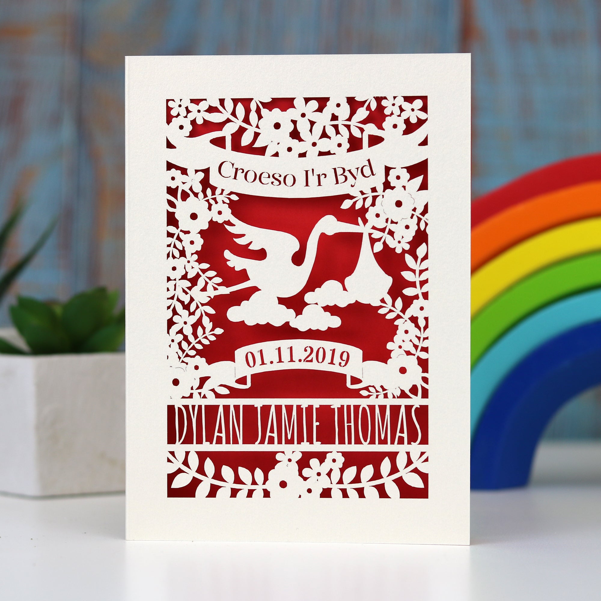 Welsh new baby personalised card. Shows Croeso i'r Byd, welcome to the world in Welsh. Has a lovely scene showing a stork carrying a baby and surrounded with leaves and flowers. All laser cut from cream card with a bright red insert. Personalise with baby's name and date of birth.