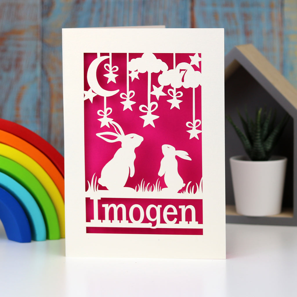 A cream and shocking pink birthday card with a name and age. Two rabbits look at the moon. 