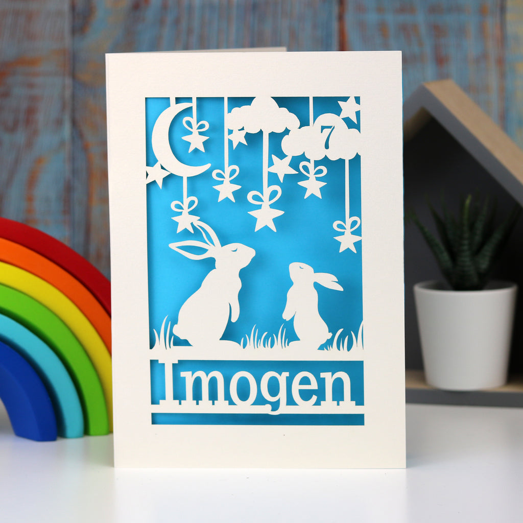 A cream and blue laser cut birthday card personalised with age and name.  Hares gaze at the moon and stars, age is laser cut from a cloud in the sky.