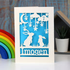 A cream and blue laser cut birthday card personalised with age and name.  Hares gaze at the moon and stars, age is laser cut from a cloud in the sky.