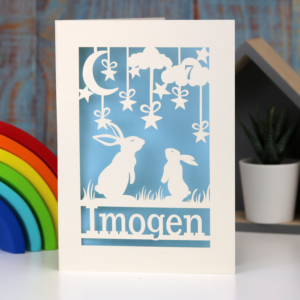 Two rabbits look up at the night sky, laser cut from a cream card and hand finished with a light blue background. Card is personalised with a name at the bottom and an age in a cloud in the sky