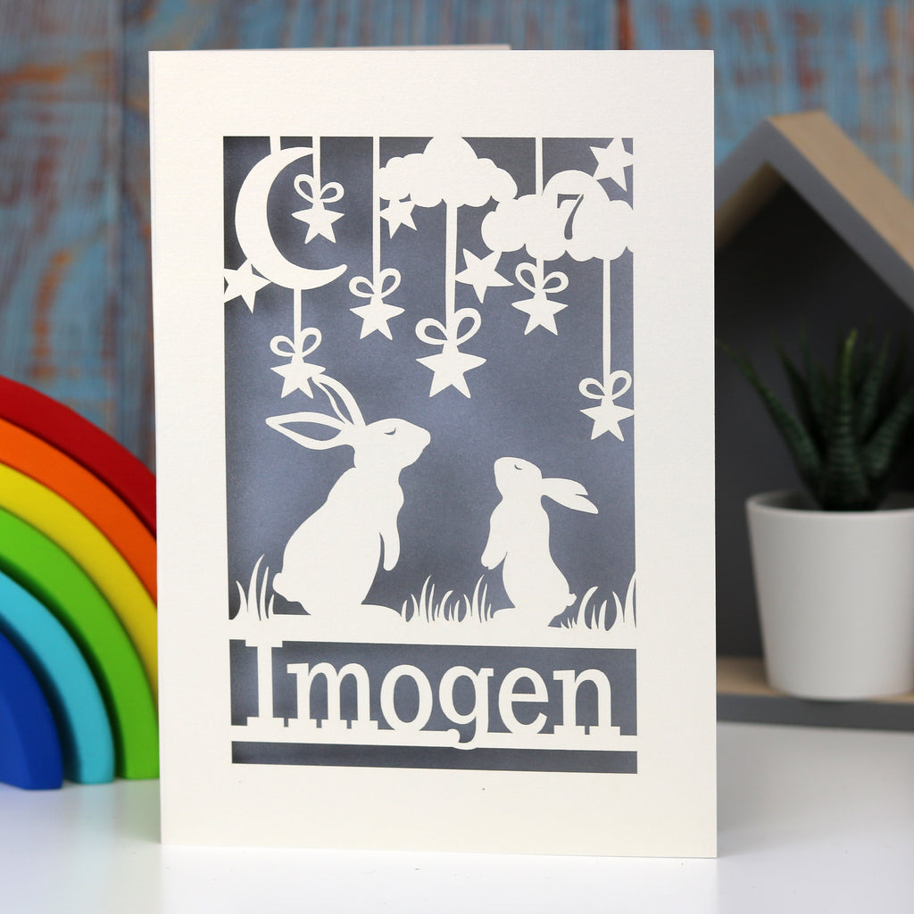 A child's birthday card laser cut from cream card with a silver paper insert. A bigger bunny and a baby bunny gaze up at the moon, with a name underneath.