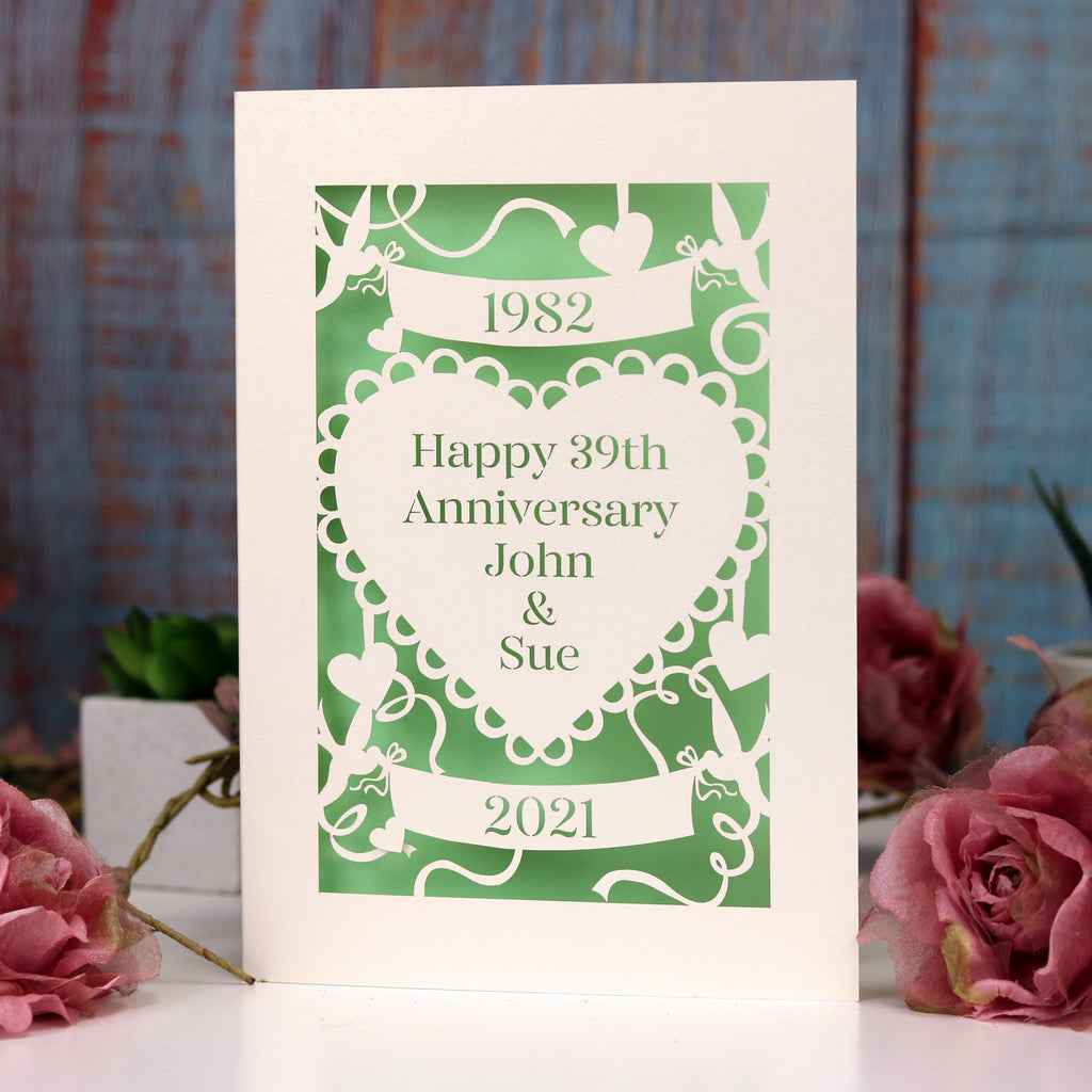 Personalised anniversary card, laser cut from cream with a light green paper behind. Design has a banner at the top with the year of the wedding, a heart in the centre with "Happy 39th anniversary John & Sue" and a banner underneath with the current year.