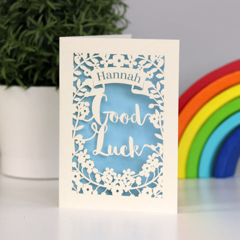 Good luck personalised laser cut cream card with a pale blue background. Shows flowers , with the words Good Luck  and a banner for the name. - A5 / Light Blue