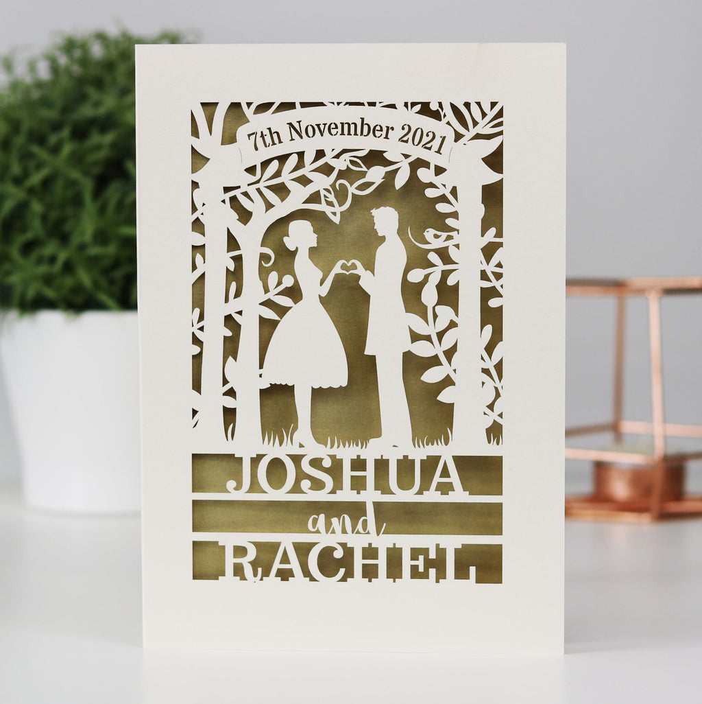A paper cut wedding card that features the silhouettes of a couple, and is personalised with a date in a banner at the top and the names of the couple over three lines of text at the bottom