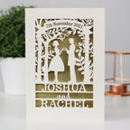 A paper cut wedding card that features the silhouettes of a couple, and is personalised with a date in a banner at the top and the names of the couple over three lines of text at the bottom