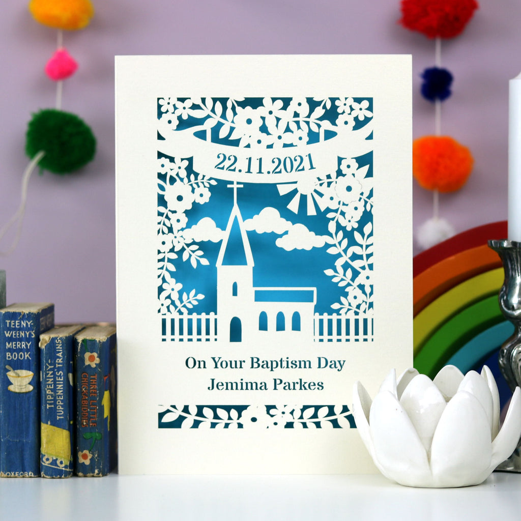 A paper cut card that reads "On Your Baptism Day" with a name underneath and a date in a banner above a cute little church. 