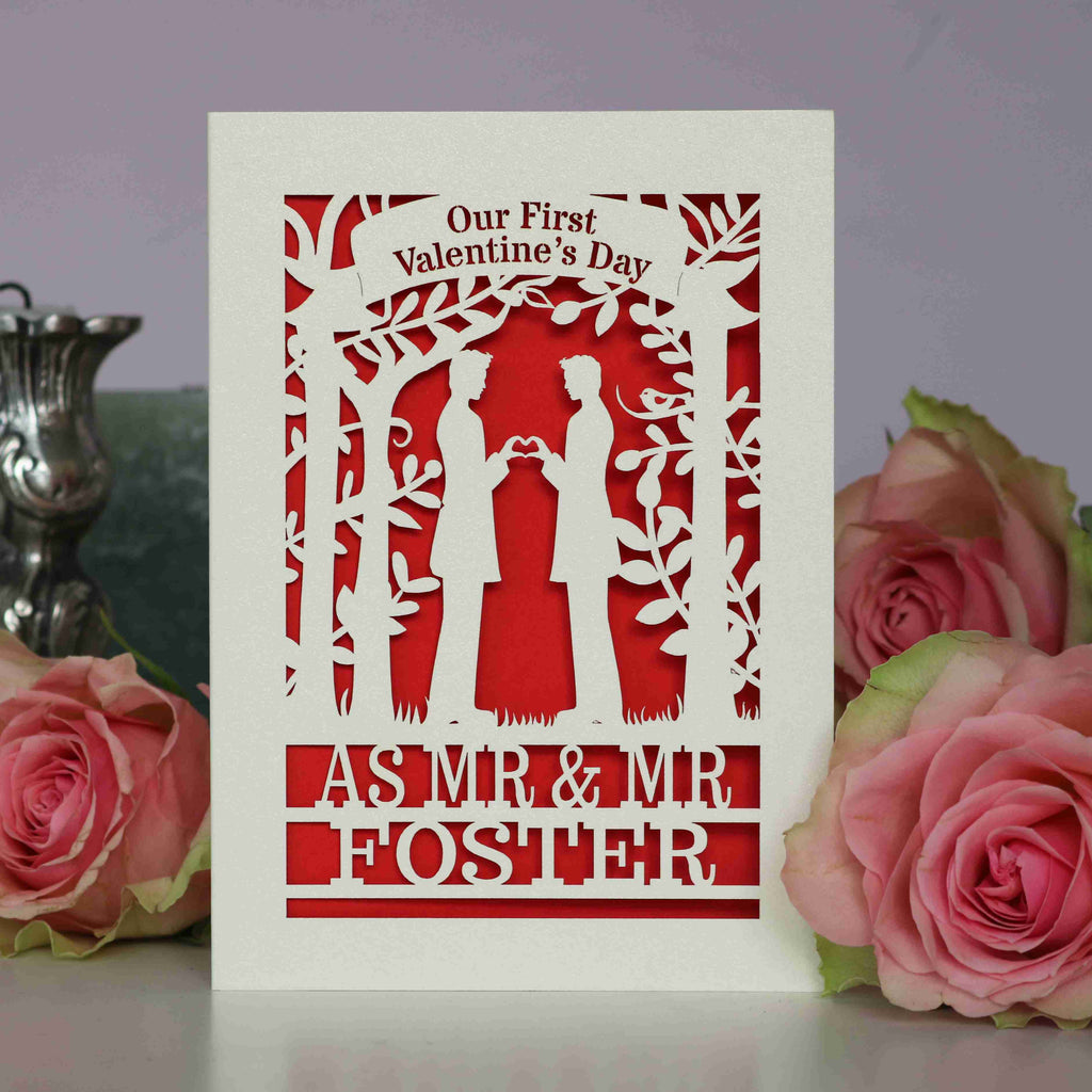 A personalised papercut gay first Valentine's card with a design of two men making a heart shape with their hands, laser cut from a cream cardstock, with a blank interior for a personal message, and a coloured paper insert visible at the bottom.