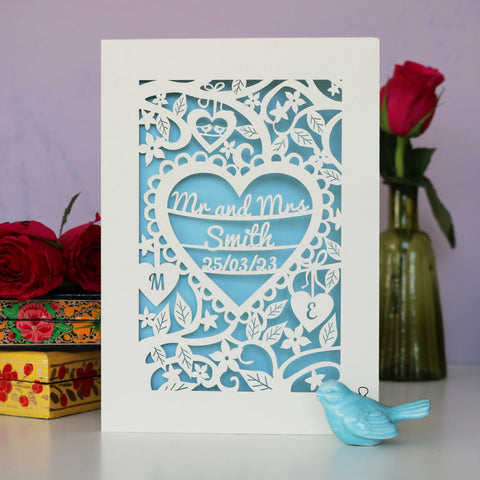 A paper cut wedding card showing names, date and initials surrounded by flowers and leaves. - A5 / Light Blue