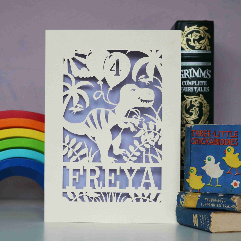 A personalised paper cut birthday card with a dinosaur on it. Dinosaur is holding balloons with an age on them and is standing on a line of text with the recipient's name cut out. 