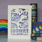 A personalised paper cut birthday card with a dinosaur on it. Dinosaur is holding balloons with an age on them and is standing on a line of text with the recipient's name cut out. 