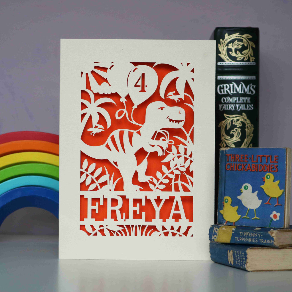 A birthday card with a dinosaur holding balloons. Card is cream with orange paper insert, and card is cut out to reveal orange behind. 