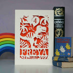 A birthday card with a dinosaur holding balloons. Card is cream with orange paper insert, and card is cut out to reveal orange behind. 