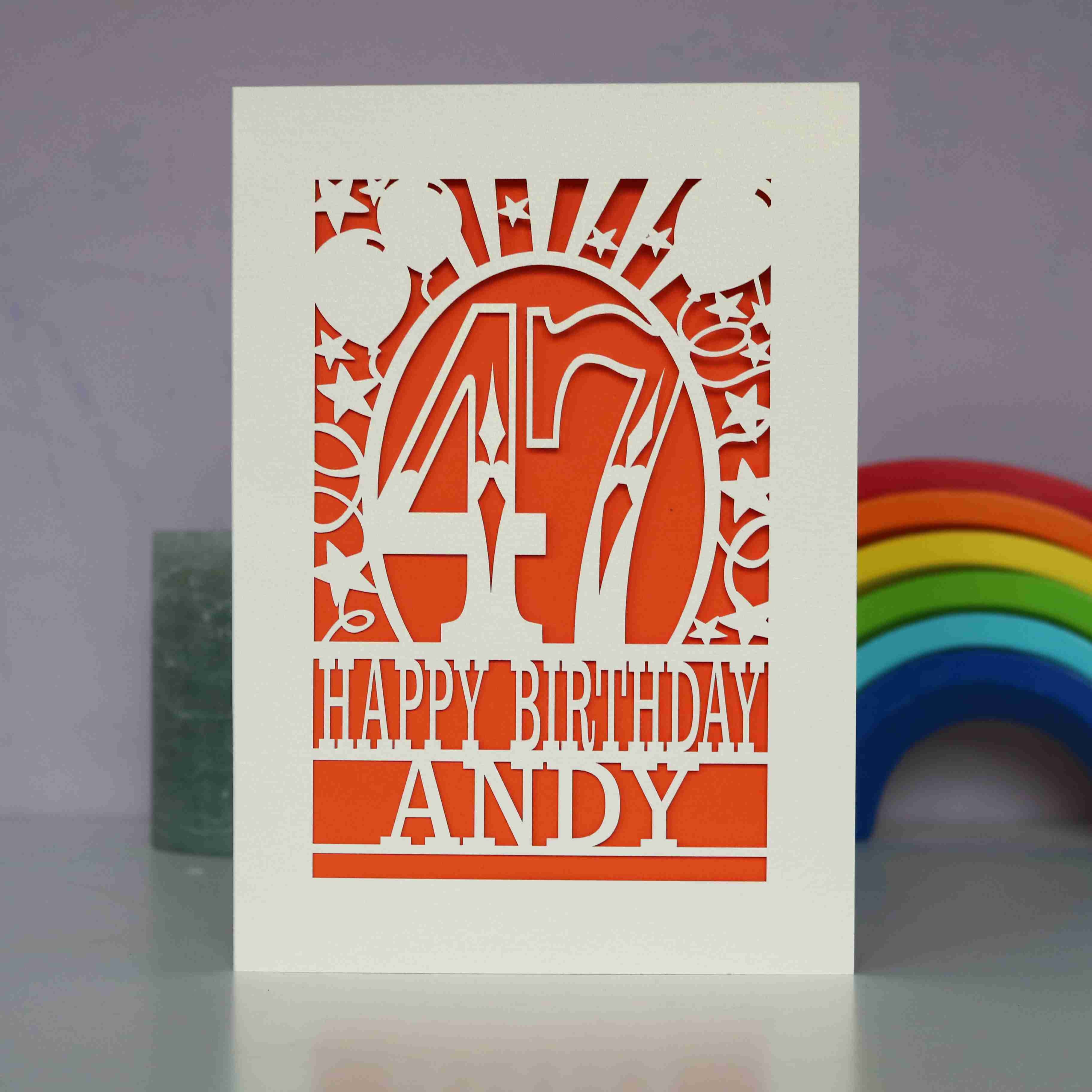 A cream paper cut birthday card cut away to reveal an orange background. Cut design is an age surrounded by stars and balloons, with the words "Happy birthday" and personalised with a name