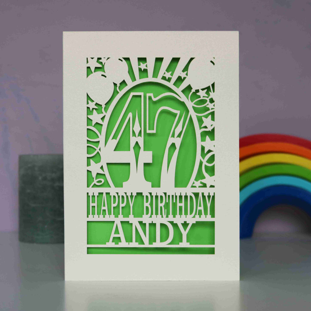 A cream paper cut birthday card cut away to reveal a green background. Cut design is an age surrounded by stars and balloons, with the words "Happy birthday" and a name underneath