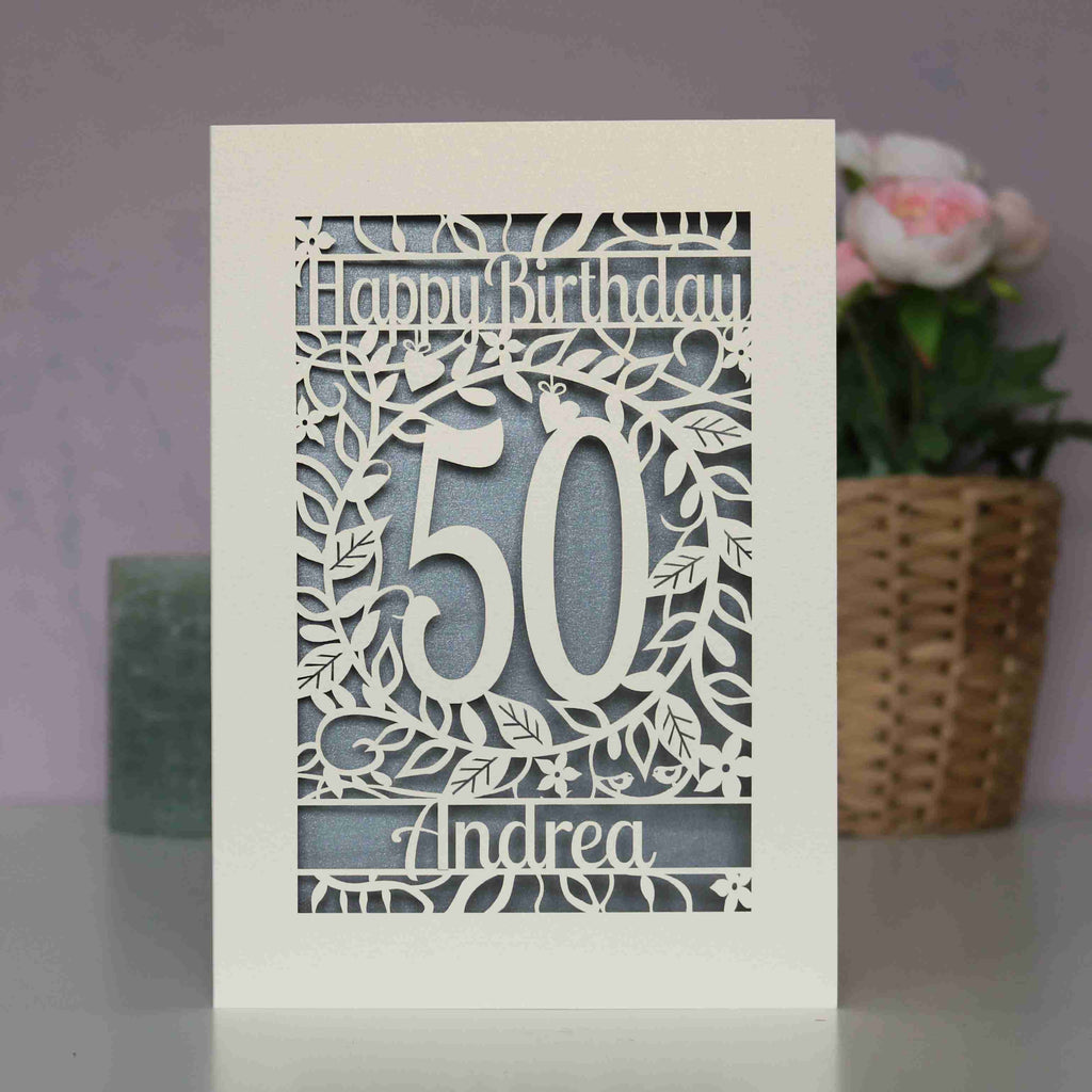 A cream and silver papercut birthday card with flowers and leaves. Card says Happy Birthday, there is an age in the middle and a name underneath.