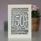 A cream and silver papercut birthday card with flowers and leaves. Card says Happy Birthday, there is an age in the middle and a name underneath.