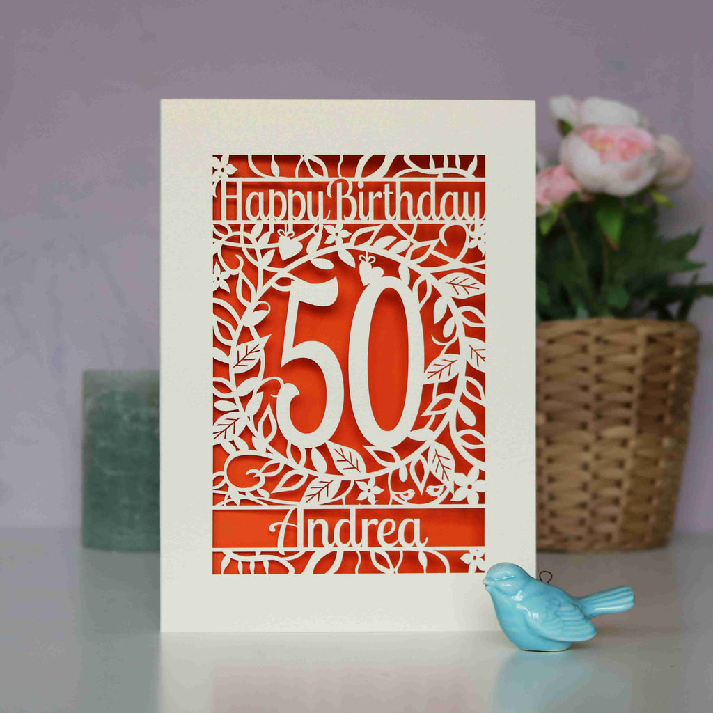 A paper cut birthday card in cream with cut out shapes to show flowers, leaves and happy birthday, with an age and a name underneath. Behind the cut away card is orange paper. 
