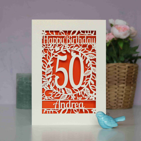 A paper cut birthday card in cream with cut out shapes to show flowers, leaves and happy birthday, with an age and a name underneath. Behind the cut away card is orange paper.  - A5 / Orange