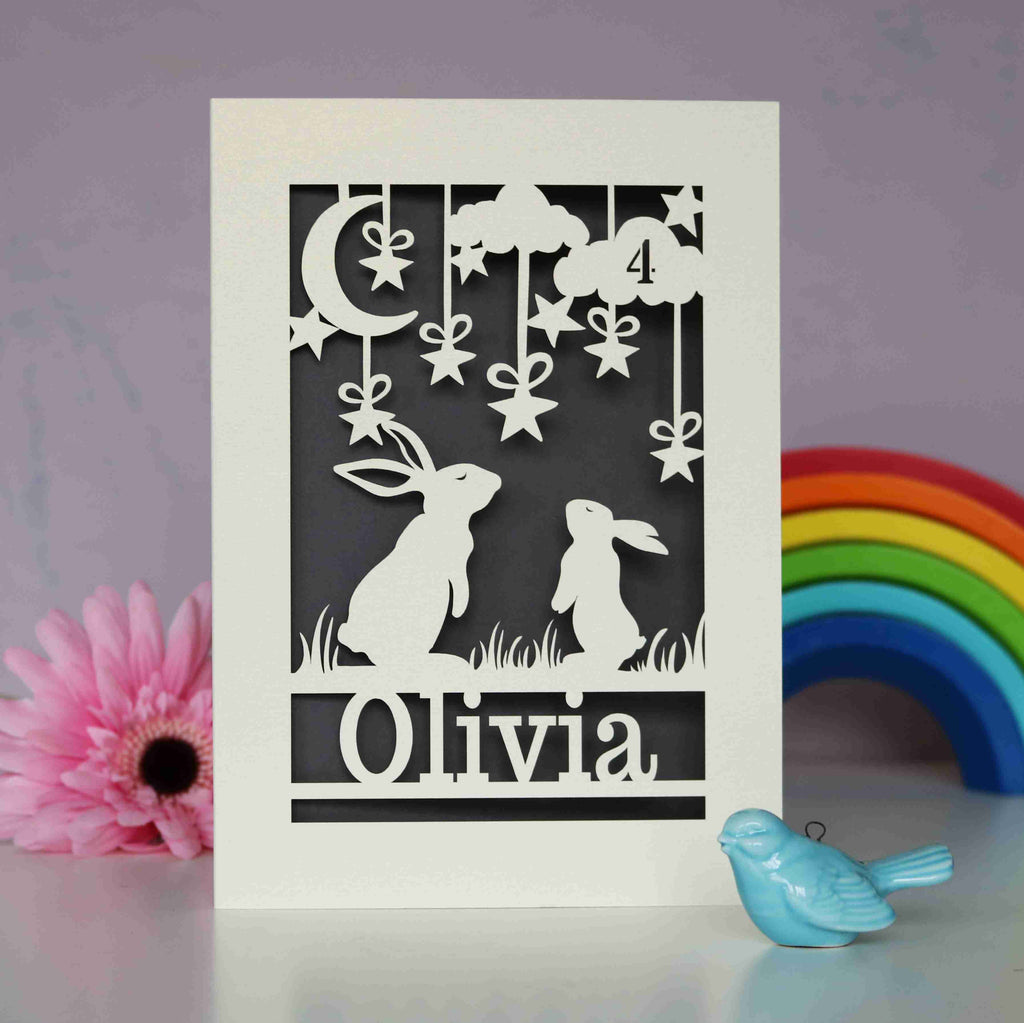 A cream and grey paper cut birthday card personalised with a name and age. Card features two bunny rabbits looking at the night sky.