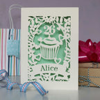 Pretty pale green cup cake personalised birthday card. Laser cut from cream card with a cupcake, an age topper, leaves, hearts and flowers. Personalise with a name and age.