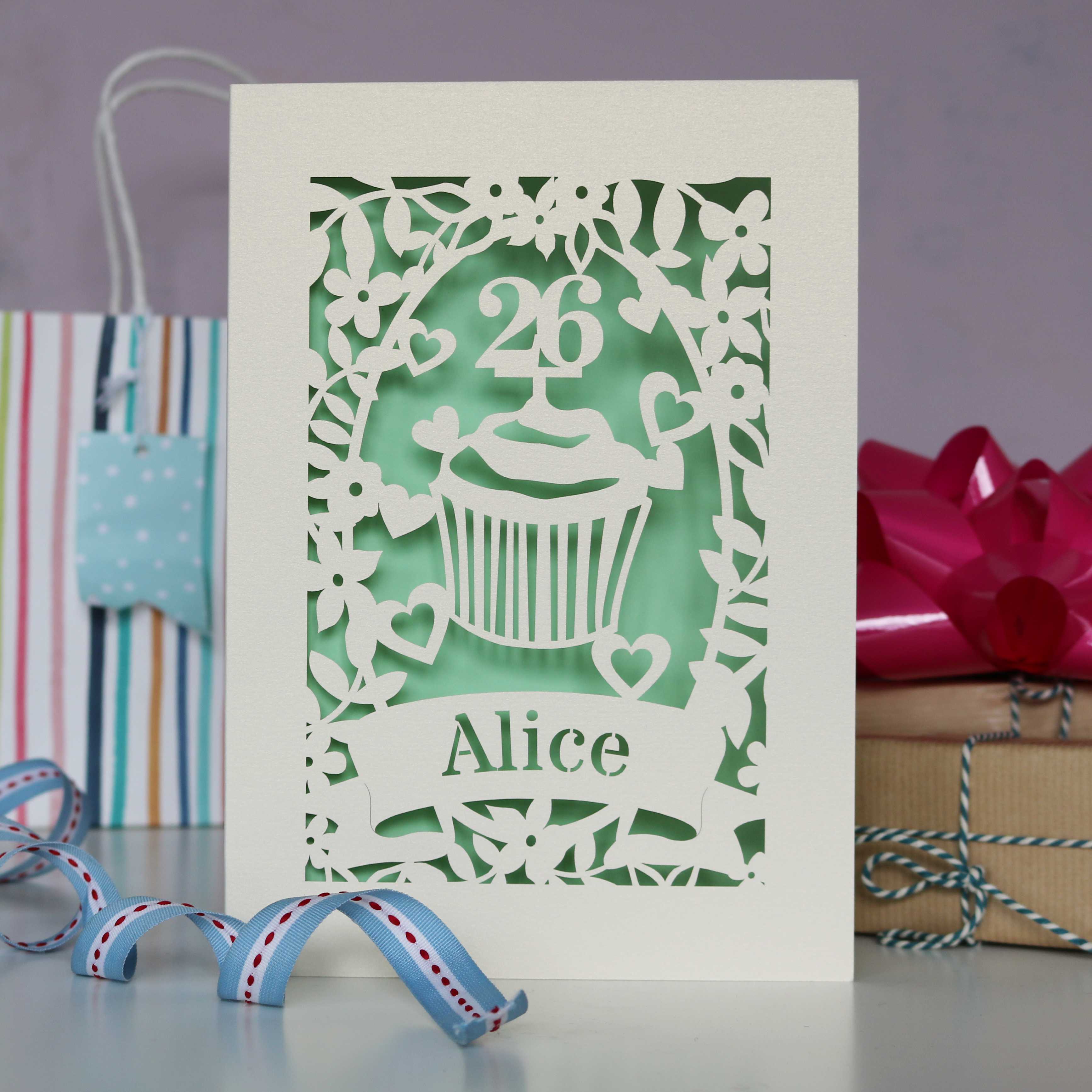 Pretty pale green cup cake personalised birthday card. Laser cut from cream card with a cupcake, an age topper, leaves, hearts and flowers. Personalise with a name and age.