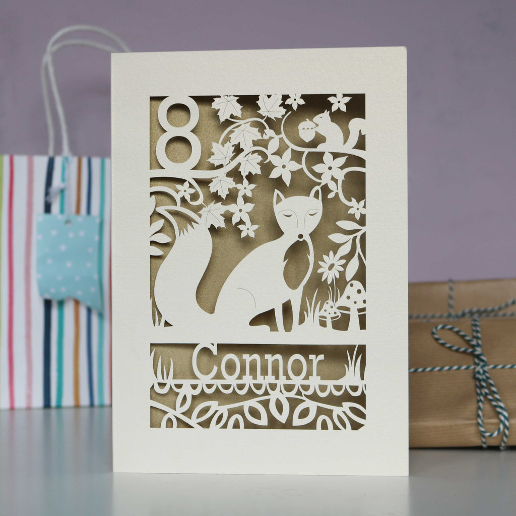 A cream and gold leaf birthday card with a fox and woodland scene. Card is personalised with an age and a name.