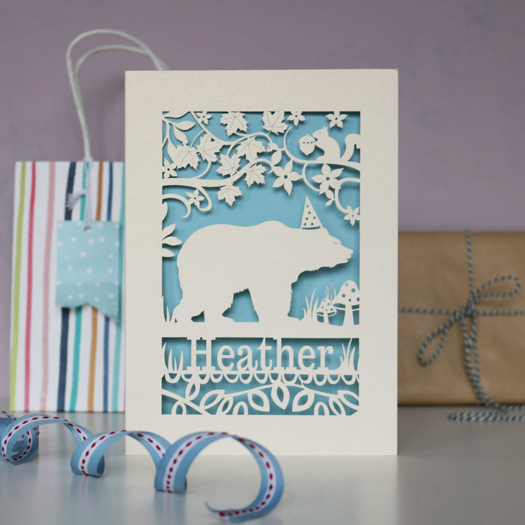 Pale blue background and cream laser cut card showing a bear in the woods.  Can be personalised with a name and optional age.
