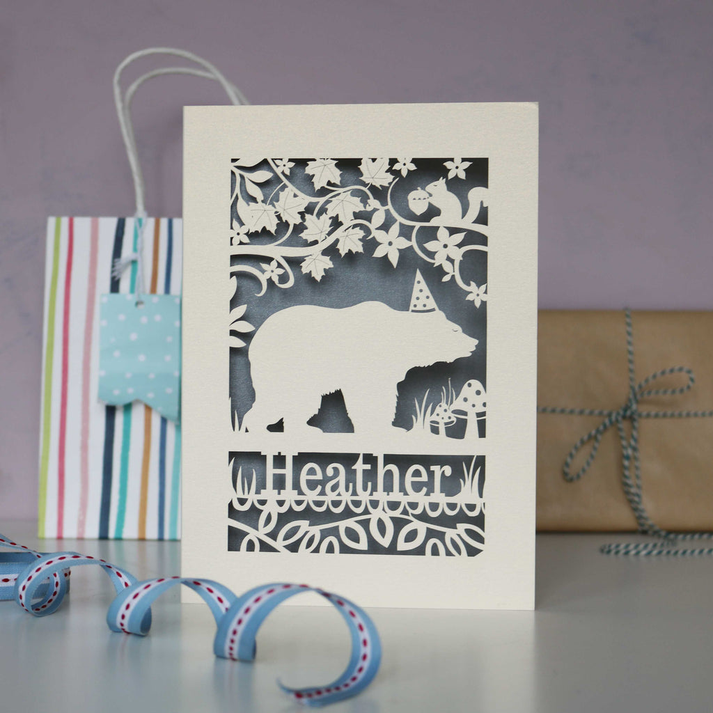 Papercut bear design birthday card.  Shows a bear wearing a hat, walking under a tree with a squirrel. Can be personalised with a name and age if you wish. Cut from cream card with a silver insert.