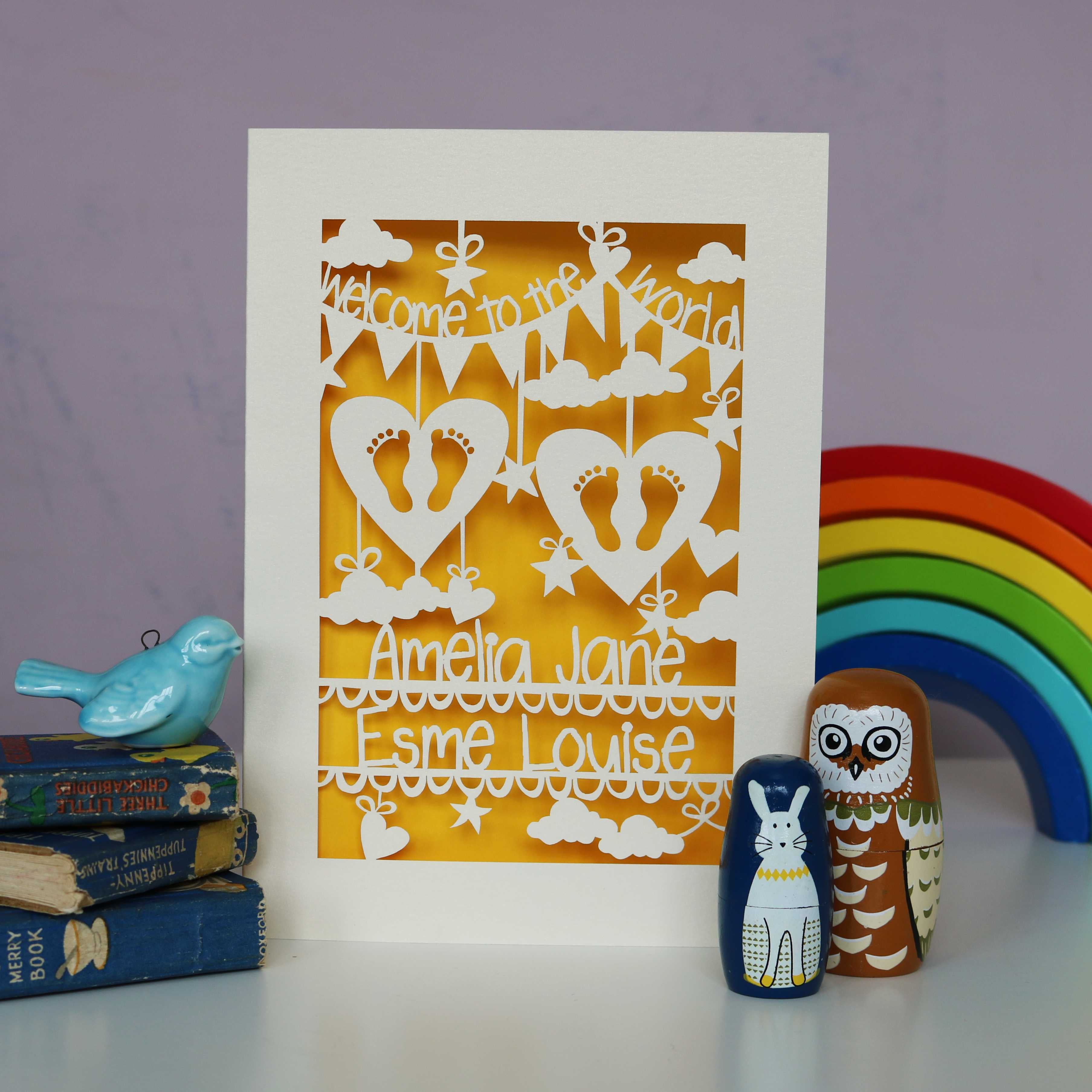 Personalised papercut card for twins, shows two sets of feet in two hearts, surrounded with clouds , stars and hearts.  Welcome to the World on bunting across the top and the babies names on two lines across the bottom. This one shown in cream card with a gender neutral sunshine yellow insert paper.