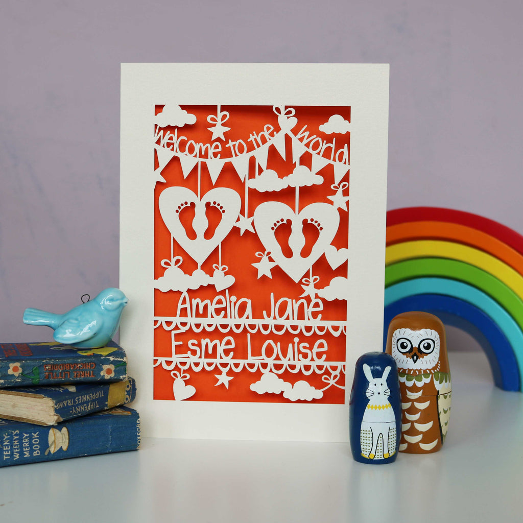 Papercut personalised twins card cut from cream card and shown with an orange background.  Space for the babies names across the bottom and "Welcome to the World" on bunting across the top. Features 2 hearts with cutout feet in both and surrounded with clouds, stars and hearts.