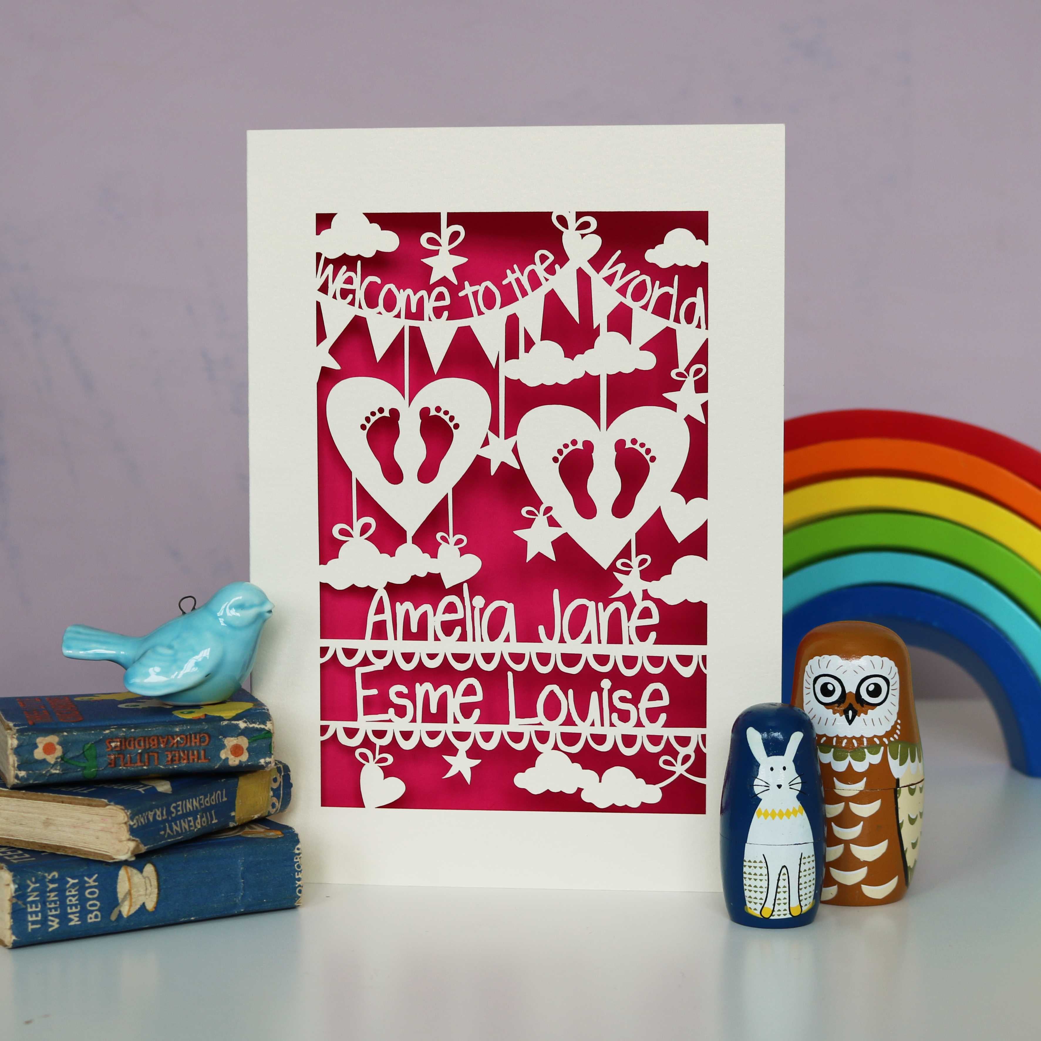 A personalised card for twin babies, personalised with their names. Card is laser cut from a cream coloured card and has a shocking pink paper insert. Card reads "Welcome to the World" and shows two sets of baby feet inside hearts, with the baby names underneath. 
