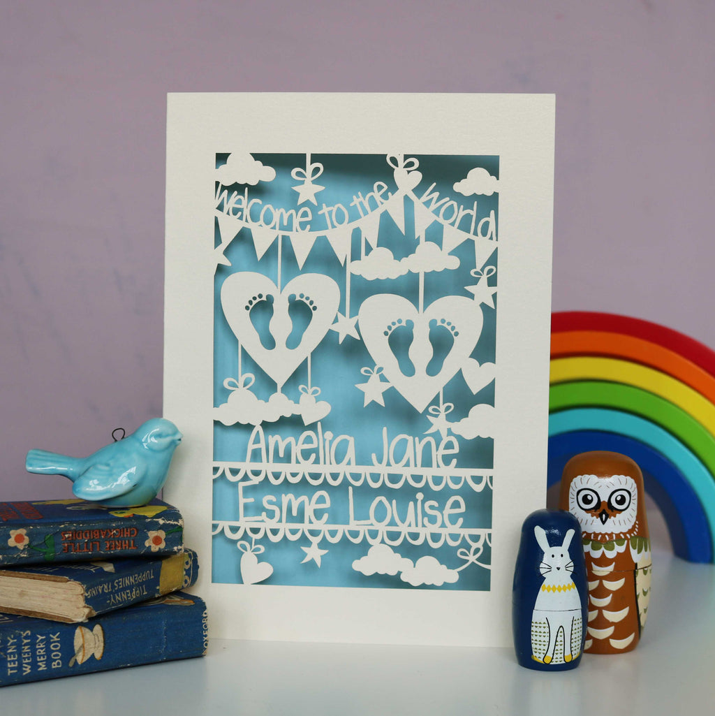 Baby blue and cream laser cut personalised twins card. Shows "Welcome to the World" in bunting across the top and the babies names across the bottom. The centre has tiny feet cut out of hearts and has clouds, stars and hearts .