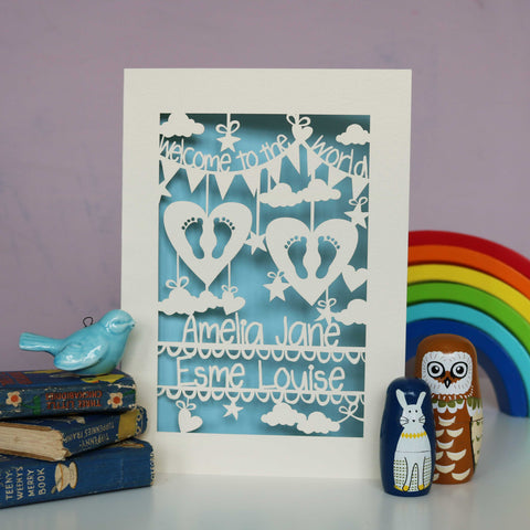 Baby blue and cream laser cut personalised twins card. Shows "Welcome to the World" in bunting across the top and the babies names across the bottom. The centre has tiny feet cut out of hearts and has clouds, stars and hearts . - A5 / Light Blue