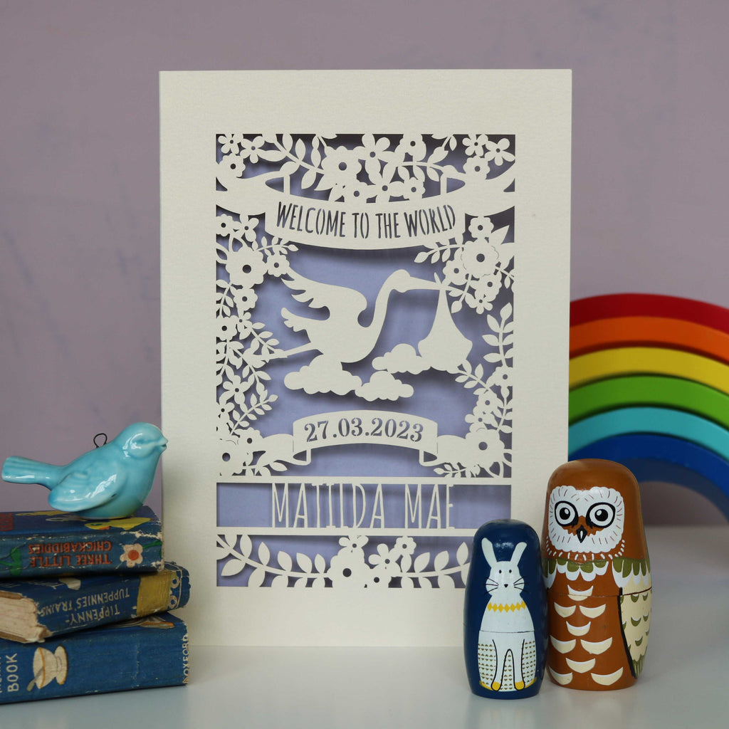 A laser cut cream and lilac new baby card personalised with the baby's name and date of birth. The top of the card reads "Welcome to the World" and the centre of the design is a stork surrounded by leaves and flowers.