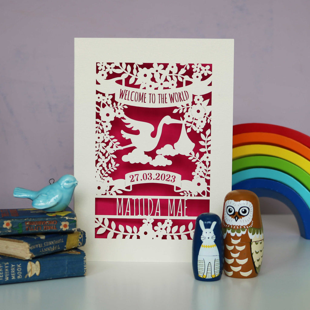 A cream new baby card with a shocking pink paper insert. The cream card is cut away to show the pink paper. Cut away design is of a stork, flowers, leaves and text. Text reads "Welcome to the world" at the top. The baby's date of birth and name are underneath the stork.