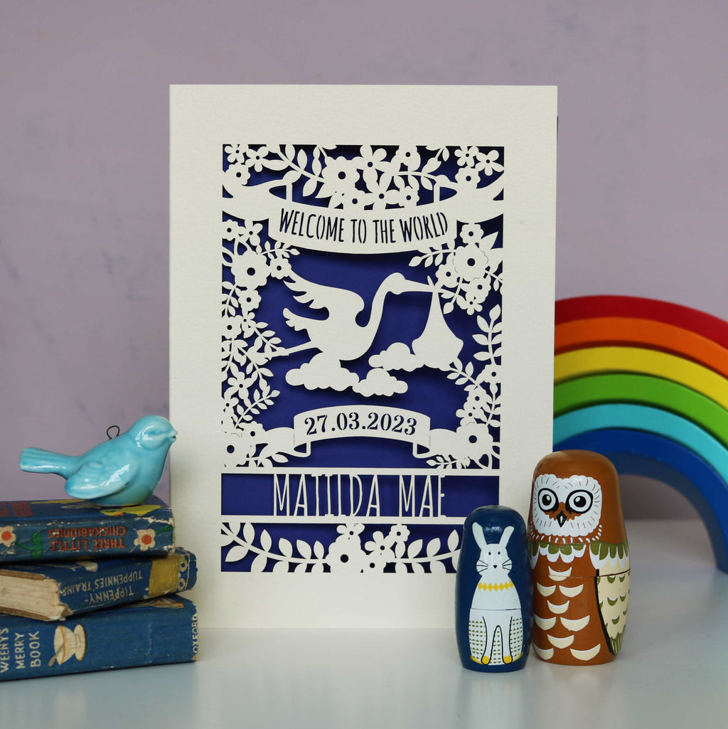 A cream new baby card with a violet coloured paper insert. The cream card is cut away to show the violet paper behind it. Cut away design is of a stork, flowers, leaves and text. Text reads "Welcome to the world" at the top. The baby's date of birth and name are underneath the stork.