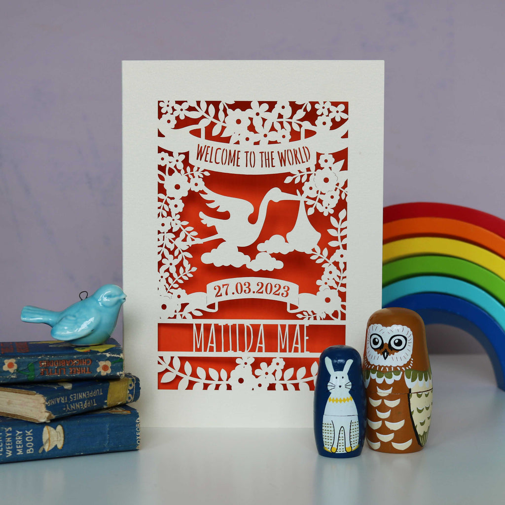 A cream new baby card with an orange paper insert. The cream card is cut away to show the orange paper. Cut away design is of a stork, flowers, leaves and text. Text r eads "Welcome to the world" at the top. The baby's date of birth and name are underneath the stork.