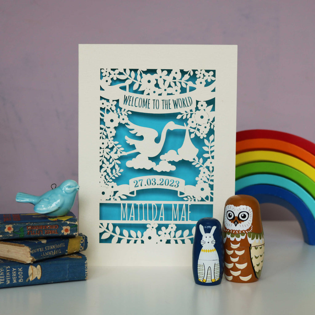 A cream and bright blue papercut card with stork in the centre, "Welcome to the World" in a banner at the top, date of birth under the stork and name at the bottom.