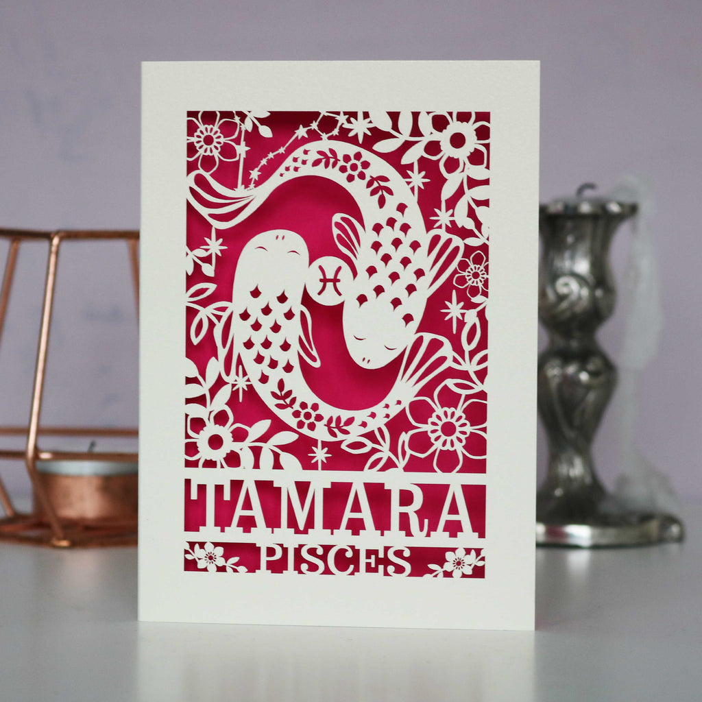 A personalised birthday card with a papercut design featuring the Pisces constellation and a fish symbol. The card is predominantly cream with a red paper cut-out in the centre, and the name 'TAMARA' is printed in large letters at the bottom.