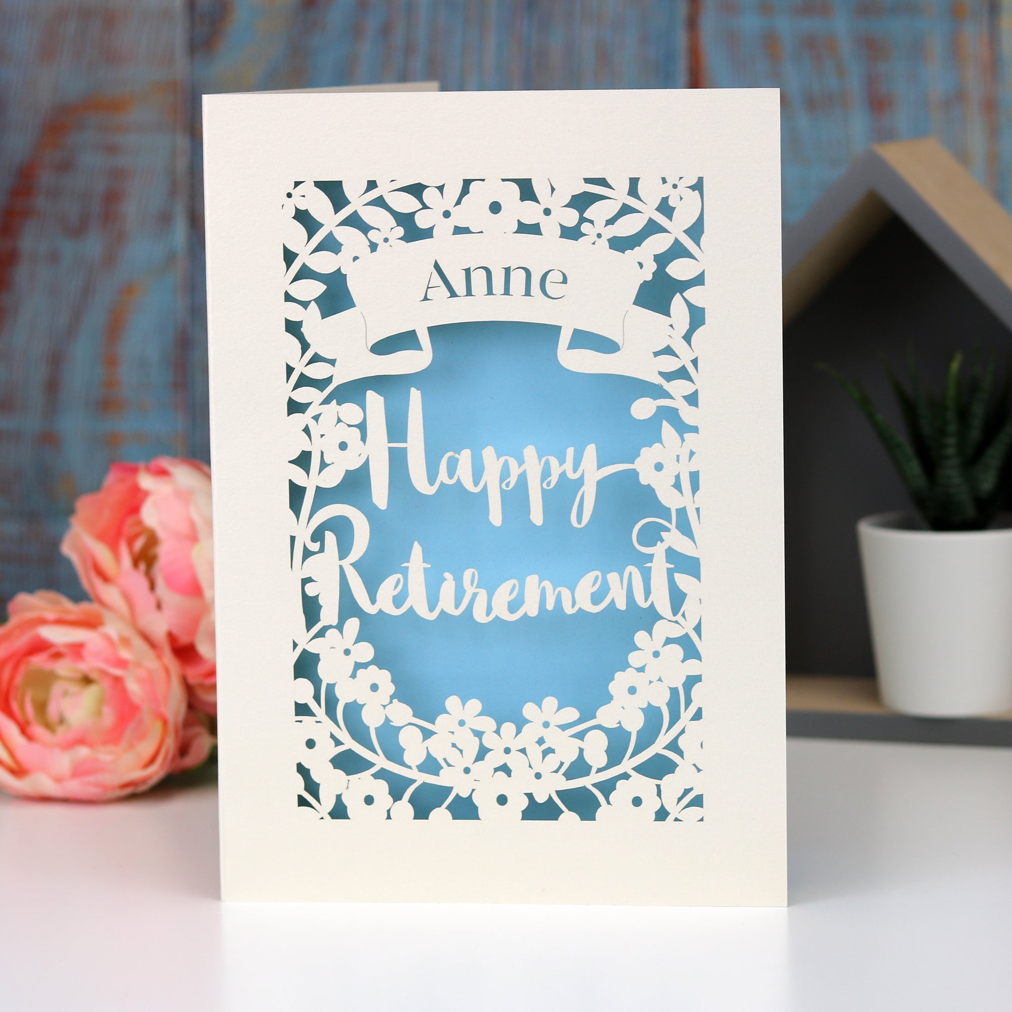 Elegant lasercut  Happy Retirement card in pale blue and cream. Shows the words Happy Retirement  surrounded with flowers  and a banner with the retiree's name,