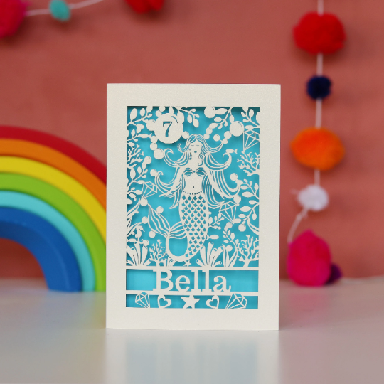 Laser cut birthday cards for children. A personalised paper cut card with a mermaid and a name and age. Card is cream with a blue paper insert. 
