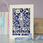 Personalised paper cut mermaids card with a name and age. Cream card and a violet paper insert behind. 