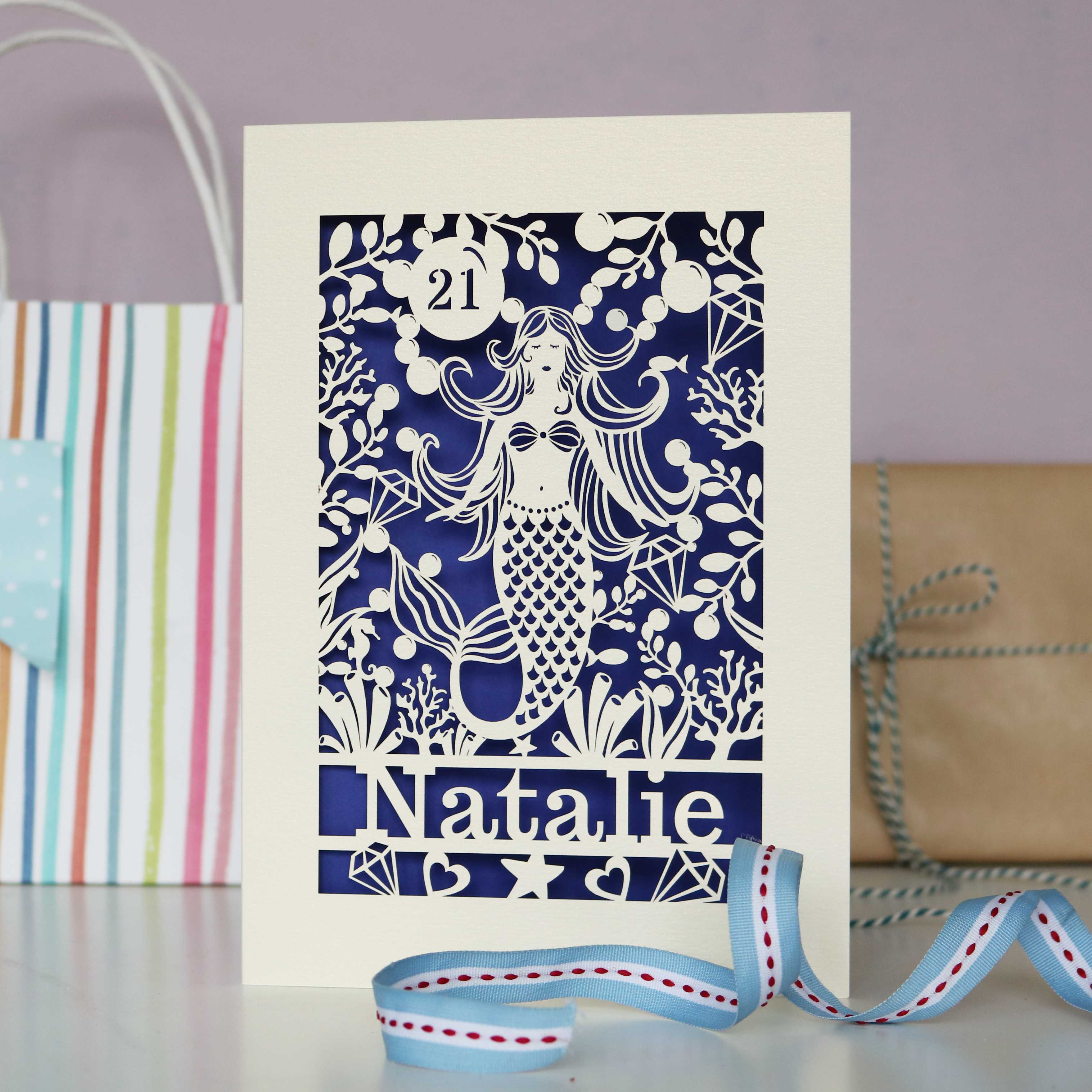 Personalised paper cut mermaids card with a name and age. Cream card and a violet paper insert behind. 